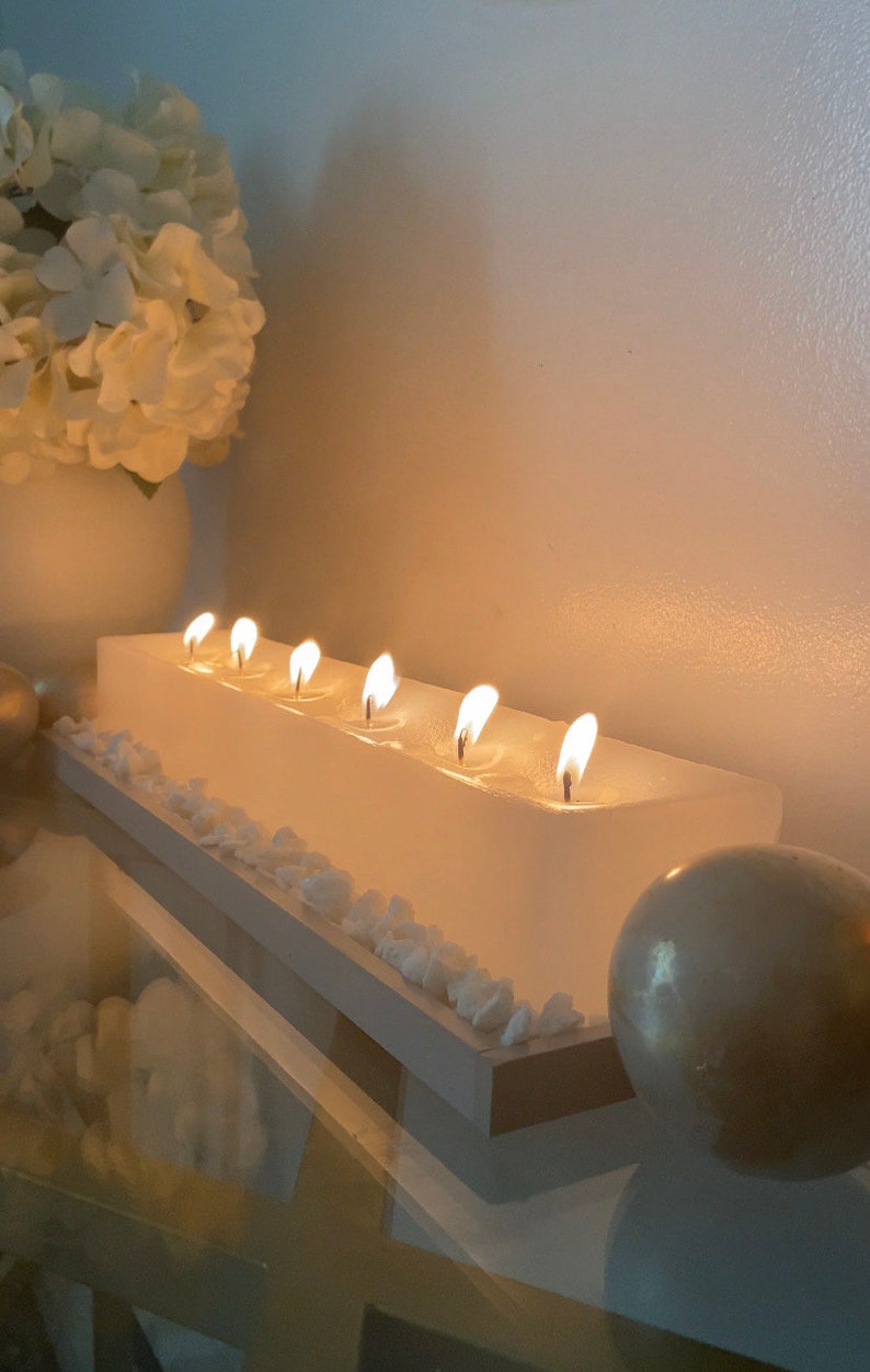 Puede incluir: Un portavelas rectangular blanco con siete velas encendidas. Las velas est&aacute;n colocadas en una base blanca, decorada con peque&ntilde;as piedras blancas. Un jarr&oacute;n blanco con flores blancas se encuentra en el fondo.
