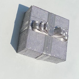 May include: A silver gift box with a silver ribbon and bow. The box is covered in small, sparkly rhinestones.