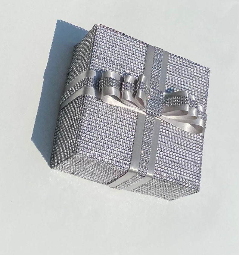 May include: A silver gift box with a silver ribbon and bow. The box is covered in small, sparkly rhinestones.