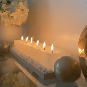 Puede incluir: Una vela blanca rectangular con seis mechas encendidas. La vela se asienta sobre una base blanca y marr&oacute;n, decorada con peque&ntilde;as piedras blancas. Un jarr&oacute;n blanco con flores blancas y esferas decorativas doradas se encuentran en el fondo.