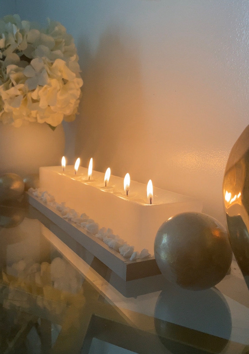 Puede incluir: Una vela blanca rectangular con seis mechas encendidas. La vela se asienta sobre una base blanca y marr&oacute;n, decorada con peque&ntilde;as piedras blancas. Un jarr&oacute;n blanco con flores blancas y esferas decorativas doradas se encuentran en el fondo.