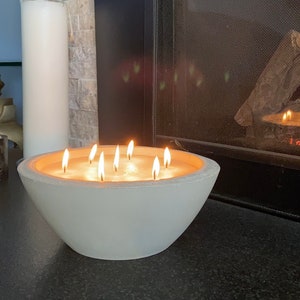 May include: A white concrete bowl with multiple lit candles inside. The bowl is sitting on a black surface in front of a fireplace.