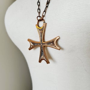 Puede incluir: Un collar con un colgante de cruz de color dorado y un diseño detallado. La cruz es grande y tiene una superficie texturizada. La cadena es de metal y tiene un diseño simple.