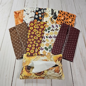 Könnte beinhalten: Ein Set mit 10 Stoff-Taschentuchbox-Bezügen in verschiedenen Herbst- und Halloween-Designs. Die Bezüge sind aus Baumwollstoff und zeigen eine Vielzahl von Motiven, darunter Sonnenblumen, Kürbisse, Fledermäuse und Blätter. Die Bezüge sind alle in verschiedenen Farben, darunter Braun, Orange, Rot und Schwarz. Die Bezüge sind auf einer weißen Holzoberfläche abgebildet.
