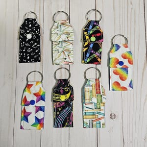 May include: Seven different keychains with various colorful patterns. The patterns include musical notes, books, crayons, and rainbows. Each keychain has a silver ring.