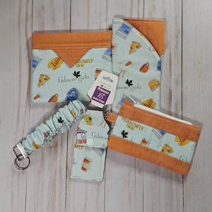 May include: A collection of fabric accessories featuring a light blue background with a repeating pattern of food, coffee cups, and the text Gilmore Girls and Dragonfly Inn, accented with orange fabric trim, including a pouch, a sleeve, and a keychain.