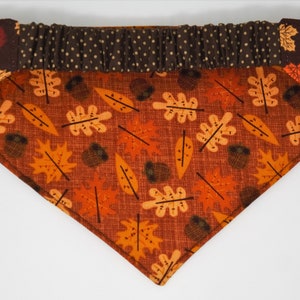 May include: A triangular pet bandana with a fall theme. The bandana features a pattern of orange and yellow leaves and acorns on a rust-colored background. The top section is brown with white polka dots, and the corners are dark brown with leaf designs.