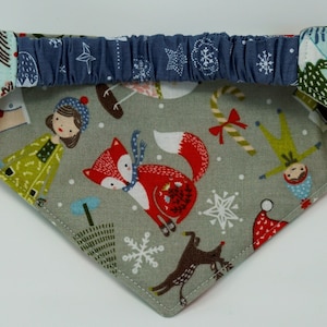 May include: A festive pet bandana with a gray background featuring a red fox, snowflakes, and holiday figures. The bandana has a blue elastic band and is trimmed with patterned fabric in shades of red, green, and blue.