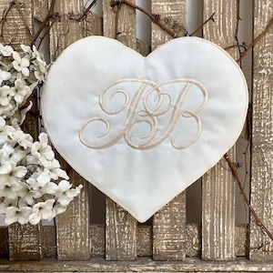 Peut inclure: Un coussin en tissu blanc en forme de cœur avec les initiales brodées "JB" en couleur crème.