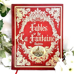 Peut inclure: Un livre à couverture rigide rouge intitulé Fables de La Fontaine illustrées avec des bordures dorées ornées et une illustration circulaire centrale représentant un loup dans un champ. Le texte Livre Deuxième est imprimé en bas.
