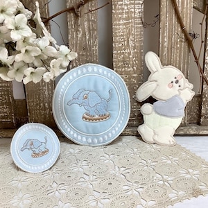 Peut inclure: Deux cercles en tissu bleu et blanc brodés avec un motif d'éléphant. Un lapin en tissu blanc avec une chemise bleue est à droite.