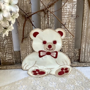 Peut inclure: Applique en forme d'ours en peluche brodée de blanc avec des accents rouges. L'ours porte un nœud papillon rouge et a des empreintes de pattes rouges sur ses pieds.