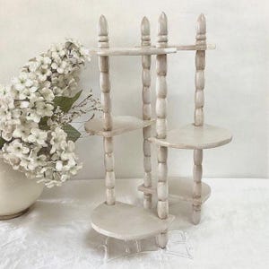 Peut inclure: Une étagère en bois à trois niveaux, de couleur blanc cassé et vieillie. L'étagère présente un design à broches avec des supports arrondis et trois étagères circulaires. Le haut de l'étagère comporte trois ornements décoratifs. Un vase de fleurs blanches est en arrière-plan.