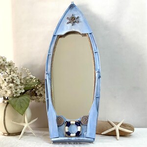 Peut inclure: Miroir en forme de bateau bleu clair, sur le thème nautique. Le miroir est orné d'une roue de navire décorative en haut et d'une bouée de sauvetage en bas. Des détails de corde et des rames complètent le design maritime. Le miroir mesure environ 91 cm de haut.