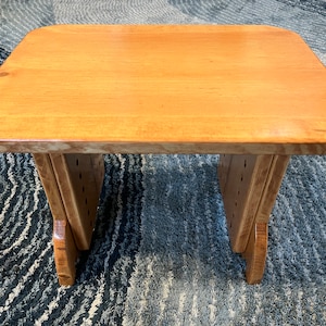 May include: A wooden stool with a natural finish and rounded edges. The stool has a simple design with two legs on each side. The stool is sitting on a patterned rug.