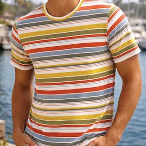 Può includere: T-shirt a maniche corte a righe orizzontali in vari colori, tra cui rosso, giallo, blu e verde. La maglietta ha un colletto bianco e viene indossata con pantaloncini beige. Lo sfondo mostra un porto turistico con barche e palme.