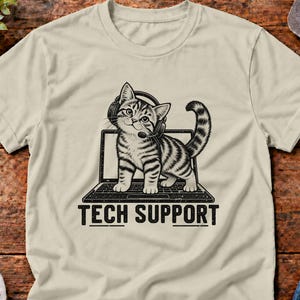 May include: A beige short-sleeve t-shirt featuring a black graphic of a tabby cat wearing a headset while standing on a laptop. Below the laptop, the text TECH SUPPORT is printed in a bold, black, sans-serif font.