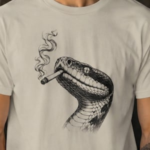 以下が含まれることがあります： ベージュ色のクルーネックTシャツ。口にタバコをくわえたヘビのモノクロのインク画風イラストが描かれており、タバコからは細い煙が立ち上っています。グラフィックは衣服の胸の中央に配置されています。