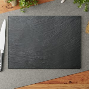 May include: A rectangular black slate serving board with a textured surface. A stainless steel knife with a black handle is on the left. The board is on a gray surface with wood and greenery in the background.