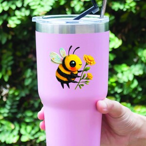 Adorable die-cut vinyl design of a fuzzy yellow honeybee holding a bright yellow flower. Shown as a fridge magnet displayed on a stainless steel refrigerator. This design is also available as a waterproof transparent sticker in five sizes. Ideal for decorating laptops, journals, water bottles, fridges, and more. Suitable for indoor and outdoor use.