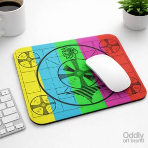 May include: A rectangular mouse mat with a colourful test pattern design. The mat features a central circular graphic with a Native American headdress and concentric circles. The background is divided into yellow, blue, green, pink, and red sections.