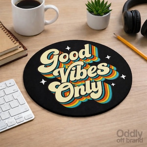 Peut inclure: Tapis de souris rond noir avec l'inscription "Good Vibes Only" dans un style rétro avec des accents arc-en-ciel. Le tapis de souris est posé sur un bureau en bois avec une tasse à café, une petite plante, un casque, un crayon et un clavier.