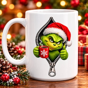 May include: A white ceramic mug featuring a graphic of a green character wearing a red Santa hat, peeking through a black zipper design. The character holds a small red mug with a white snowflake pattern, while giving a thumbs up gesture.