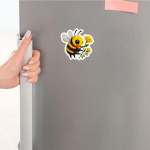Adorable die-cut vinyl design of a fuzzy yellow honeybee holding a bright yellow flower. Shown as a fridge magnet displayed on a stainless steel refrigerator. This design is also available as a waterproof transparent sticker in five sizes. Ideal for decorating laptops, journals, water bottles, fridges, and more. Suitable for indoor and outdoor use.