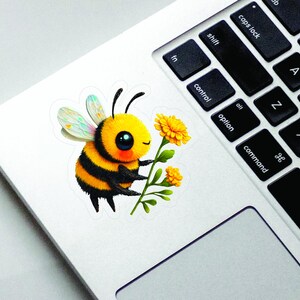 Adorable die-cut vinyl design of a fuzzy yellow honeybee holding a bright yellow flower. Shown as a fridge magnet displayed on a stainless steel refrigerator. This design is also available as a waterproof transparent sticker in five sizes. Ideal for decorating laptops, journals, water bottles, fridges, and more. Suitable for indoor and outdoor use.