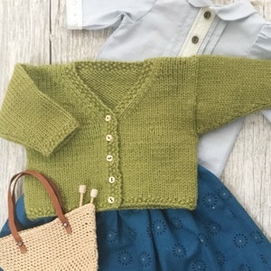 Peut inclure: Un cardigan en tricot vert avec un col en V et une fermeture à boutons. Le cardigan est posé sur un tissu à motifs bleus et blancs.