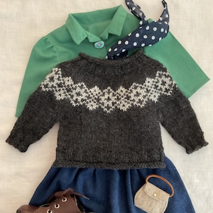 May include: A dark gray knitted sweater with a white geometric pattern around the neckline. The sweater is worn over a green shirt with a white collar and a black and white polka dot bow tie. The sweater is paired with a dark blue skirt and a small brown purse.