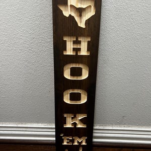 May include: A vertical wooden sign with a dark brown stain. The top features a cutout of the state of Texas with a longhorn. Below, the sign reads "HOOK 'EM" in large, carved letters, with a hand gesture at the bottom.