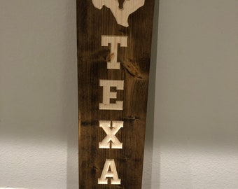 May include: A wooden sign with the word "TEXAS" carved in white letters. The state of Texas is carved above the word, with a longhorn steer inside the state outline.