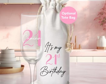 May include: A white tote bag with the text "It's my 21st Birthday" in pink and black lettering. A champagne flute with the number 21 and the name "Cameron" in pink lettering is next to the bag. The bag is optional.