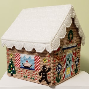 May include: A white gingerbread house tissue box cover with a brown base and colorful candy cane and gingerbread details. The roof is covered in white felt and has a scalloped edge.