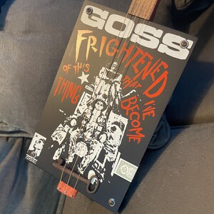 May include: A handmade cigar box guitar with a black body featuring the text "GOSS FRIGHTENED OF THIS THING THAT WE'VE BECOME" in white and red. The guitar has a wooden neck and strings.