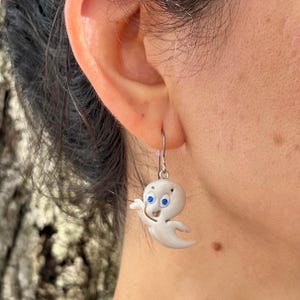 May include: A single white ghost-shaped earring with blue eyes and a friendly expression. The earring is hanging from a silver hook. The earring is a whimsical accessory.