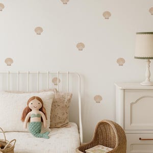 Op de afbeelding: Een kinderkamer met een wit metalen bed, een zeemeerminnenpop en een rieten wieg. De muur is versierd met schelpenstickers. Een wit nachtkastje met een lamp staat rechts van het bed. De kamer heeft een zacht, neutraal kleurenpalet.