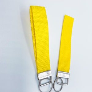 Puede incluir: Dos llaveros de muñeca amarillos brillantes. Están hechos de tela amarilla tejida, con herrajes metálicos plateados y anillas para llaves. El fondo es blanco.