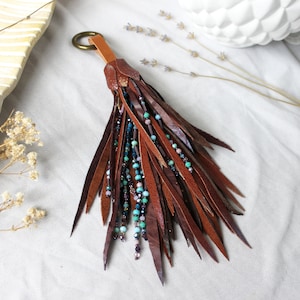 Peut inclure: Un pompon décoratif composé de nombreuses fines lanières de cuir marron et marron foncé, entremêlées de brins de petites perles multicolores dans des tons turquoise, violet et verre transparent, fixé à un anneau circulaire en laiton.