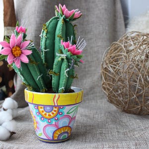 May include: A handmade felt cactus in a patterned pot. The cactus is green with pink flower details and white stitching. The pot has a yellow rim and colourful floral patterns. A decorative item for the home.
