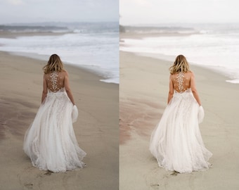 May include: A bride in a white lace wedding dress with a beaded back walks on a sandy beach with the ocean in the background.