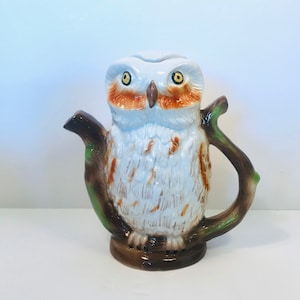 May include: A white ceramic owl teapot with brown and orange markings on its face and body. The teapot is perched on a brown branch with green leaves. The teapot has a spout and a handle.