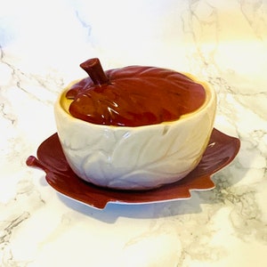 May include: A ceramic apple-shaped dish with a matching lid and leaf-shaped saucer. The lid and saucer are a deep reddish-brown, while the dish itself is off-white with leaf-like detailing. The stem of the apple is brown.