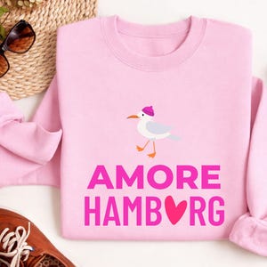 Könnte beinhalten: Hellrosa Sweatshirt mit den Worten "AMORE HAMBURG" in rosa Blockbuchstaben. Über dem Text befindet sich eine Cartoon-Möwe mit rosa Mütze. Sonnenbrille und braune Schuhe sind im Hintergrund zu sehen.