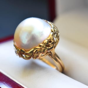 May include: A gold ring featuring a large, round, iridescent white pearl set within an intricate, ornate yellow gold mounting. The metalwork surrounding the base of the pearl displays detailed, raised floral and leaf-like scroll patterns.