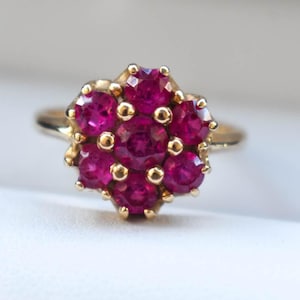 Op de afbeelding: Een gouden ring met een cluster van dieprode edelstenen gerangschikt in een bloemmotief. De ring wordt gepresenteerd in een witte doos met een rode rand, wat het ontwerp en de kleur van de ring benadrukt.