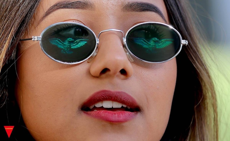 Puede incluir: Primer plano de una persona que lleva gafas de sol ovaladas con un dise&ntilde;o de p&aacute;jaro verde reflejado en las lentes. La persona lleva l&aacute;piz labial rojo y tiene el pelo oscuro.