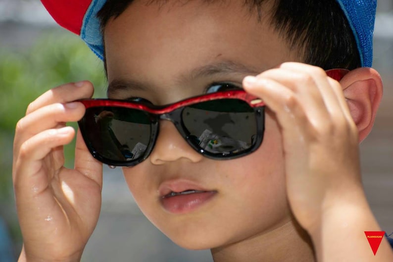 May include: A young person wearing a red and blue baseball cap and sunglasses with red frames. The sunglasses are black and have a reflective lens.