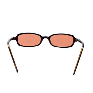 May include: Rectangular sunglasses with a dark brown tortoiseshell frame and light orange tinted lenses. The glasses feature thin, curved temples and a bridge connecting the two oval-shaped lenses. The design is simple with a smooth finish.
